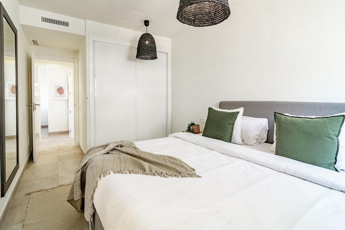 A bedroom with a bed, a bedside table, a hanging light fixture, and a door leading to another room.