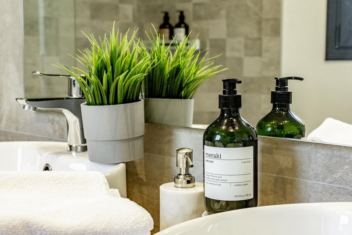 A bathroom with a sink, two green bottles labeled 'meraki', a soap dispenser, and two potted plants.