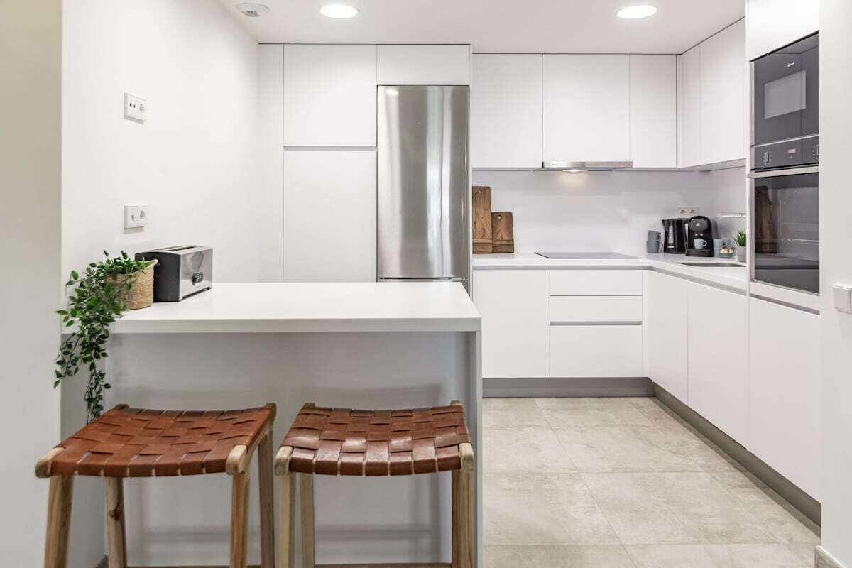 A modern kitchen with white cabinets, a stainless steel refrigerator, a built-in oven, and a small island with stools.