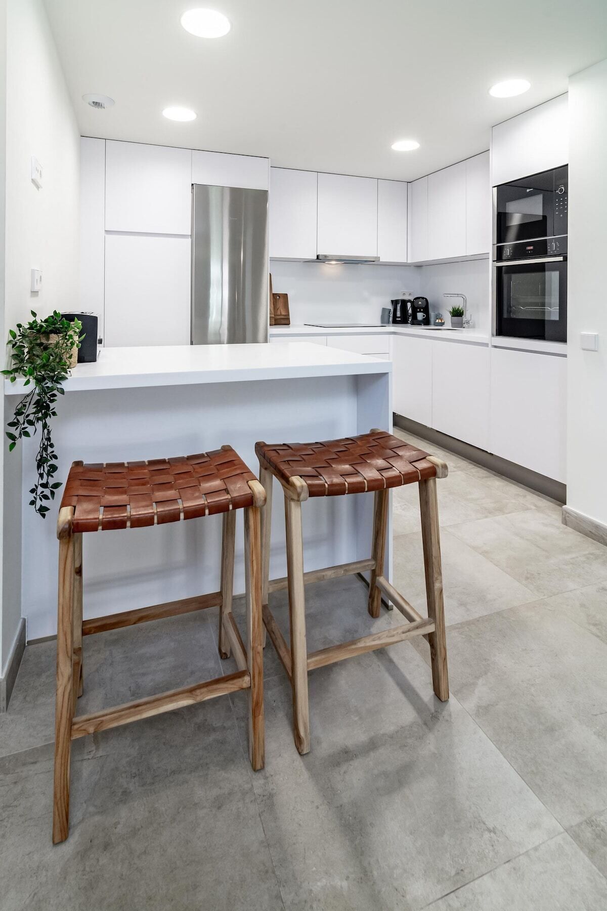 A modern kitchen with white cabinets, stainless steel appliances, and two wooden stools with woven seats.