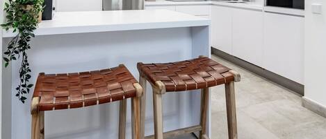 A modern kitchen with white cabinets, stainless steel appliances, and two wooden stools with woven seats.