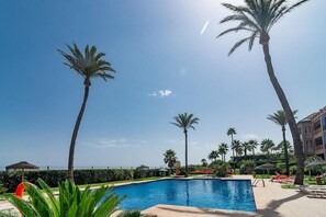 A swimming pool with palm trees, a clear sky, and a building in the background. - Lap- 3 bed Apartment on the Beach. Families Only (Casares)