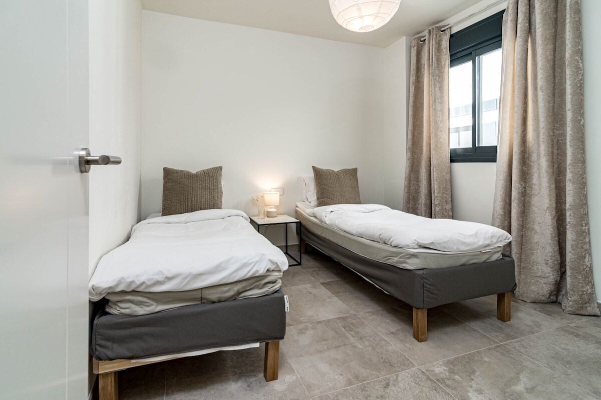 Un dormitorio pequeño y moderno con dos camas individuales, una mesita de noche con lámpara y una ventana con cortinas.