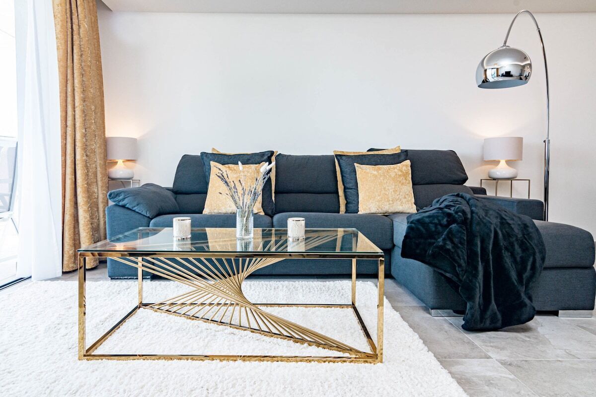 Uma sala de estar moderna com um sofá azul escuro, duas mesas laterais com abajures, uma mesa de centro de vidro com moldura dourada e uma janela grande com cortinas.