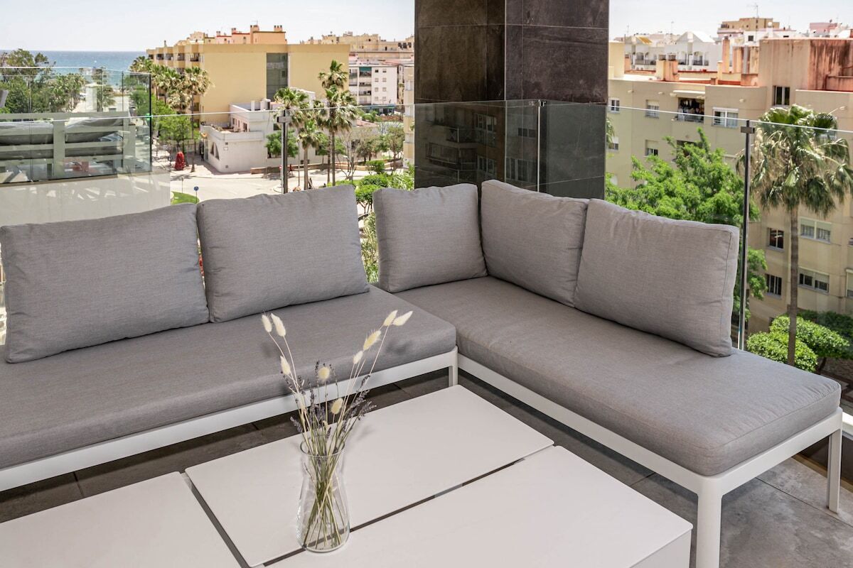 Un balcon moderne avec un canapé d’angle gris et une table basse blanche, offrant une vue sur un paysage urbain côtier, avec des palmiers et des bâtiments.