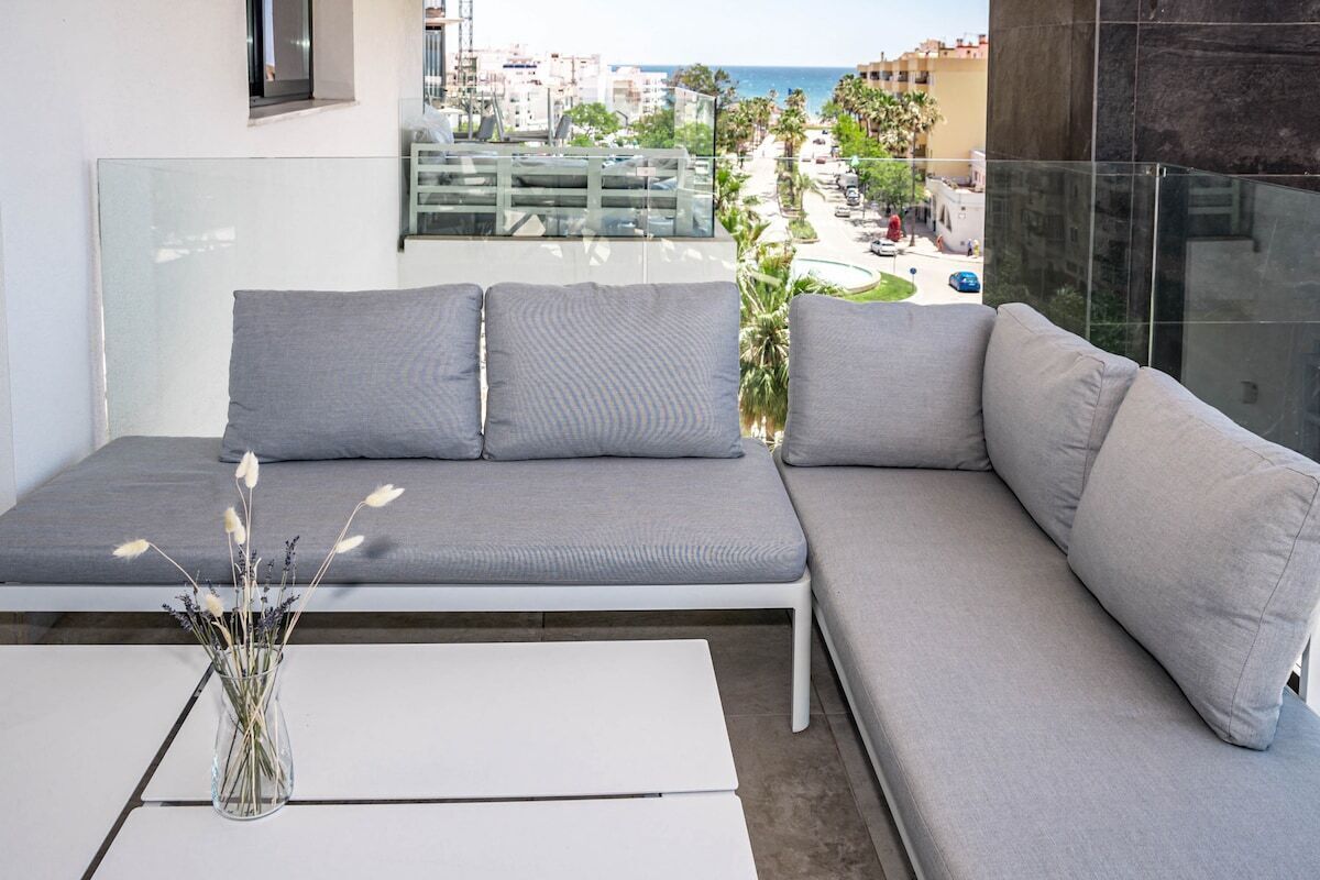 A modern balcony with a grey sofa and a white coffee table, overlooking a cityscape and the sea.