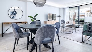A modern dining area with a round table, grey upholstered chairs, and a large window offering a view of the city. - Inf32l- Modern City Apartment, Families Only (Estepona)