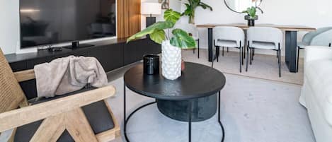 A modern living room with a flat-screen TV, a round coffee table with a plant, a sofa, and a dining area with chairs and a table.