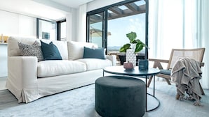 A modern living room with a white sofa, a round coffee table, and a round ottoman. There is a large window with a view of the outdoors. - Lme9f1- Penthouse, Amazing Views, Families Only (Estepona)