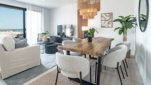 A modern living room with a wooden dining table, grey chairs, a sofa, a TV, and a large window overlooking the beach. - Lme9f1- Penthouse, Amazing Views, Families Only (Estepona)