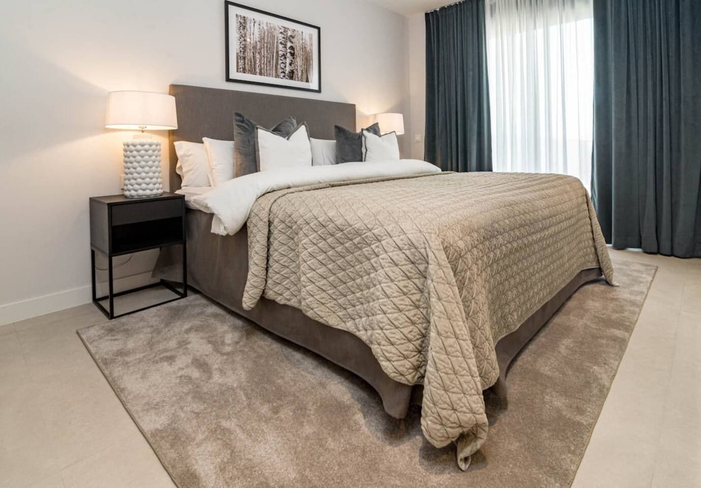 A bedroom with a bed, bedside tables, a lamp, a framed picture on the wall, and a window with curtains.