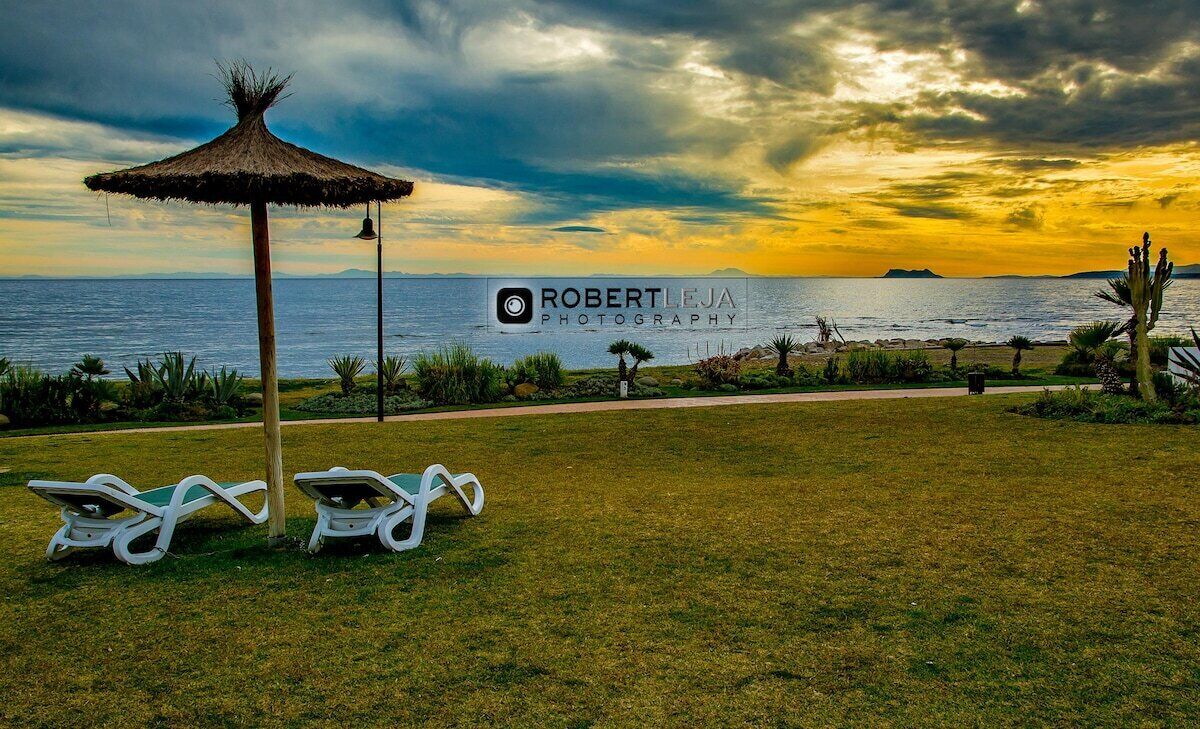 A grassy area with two white lounge chairs and a thatched umbrella, a path leading to the sea, and a logo for Robert Leja Photography.