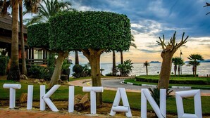 A sign with the word "TIKITANO" in large white letters, surrounded by a landscaped area with palm trees and a view of the sea. - Lm352b- Spacious Duplex Penthouse With Panoramic (Estepona)