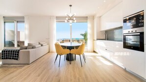 A modern living room with a sofa, dining table, and chairs, a kitchen with appliances, and a large window with a view. - Lm352b- Spacious Duplex Penthouse With Panoramic (Estepona)