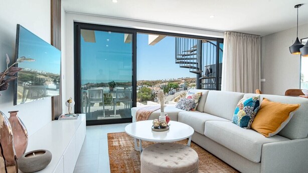 Una sala de estar moderna con un sofá, un reposapiés y una mesa de centro, ventanas amplias con vistas a la ciudad y una escalera.
