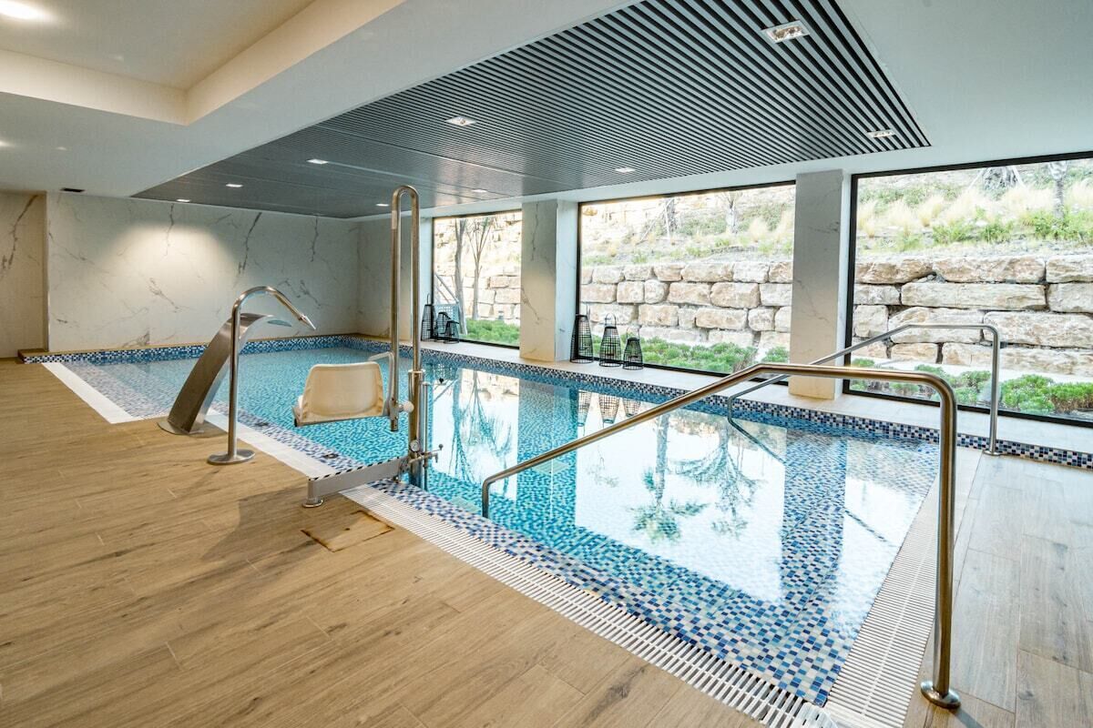 A modern indoor swimming pool with clear glass walls, a wooden floor, and a stone wall backdrop.