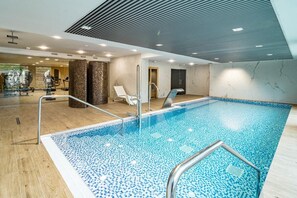 Una piscina interior con suelo de baldosas, una escalera de metal y un techo con diseño lineal.