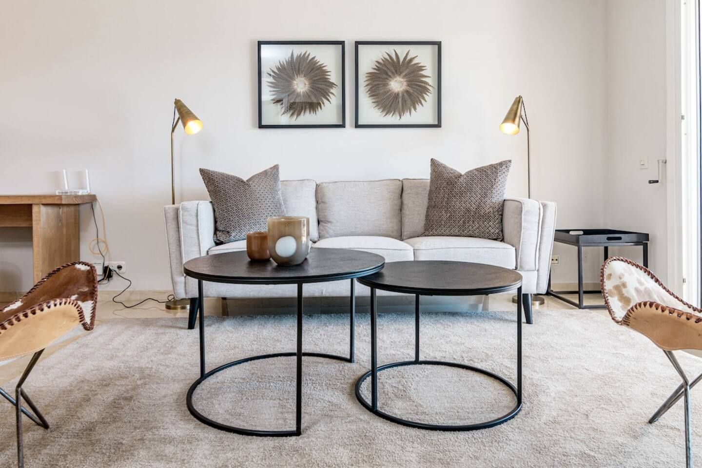 Ein modernes Wohnzimmer mit einer Couch, zwei Couchtischen und dekorativen Kissen. An der Wand befinden sich zwei gerahmte Kunstwerke und zwei Stehlampen.