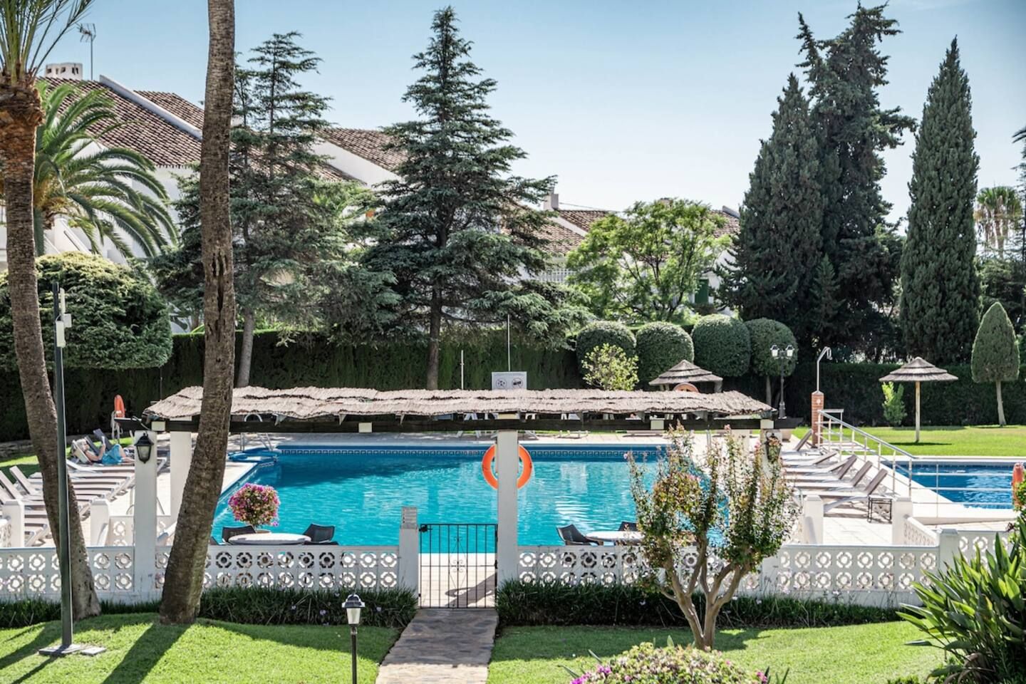 Una piscina con un deck circundante, sillas de descanso y un jardín bien cuidado con árboles y arbustos.