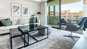 A modern living room with a glass coffee table, a white sofa, and a grey armchair. There are framed pictures on the wall and a large window offering a view of buildings. - In33k- Stunning City Apartment Close to Beaches (Estepona)