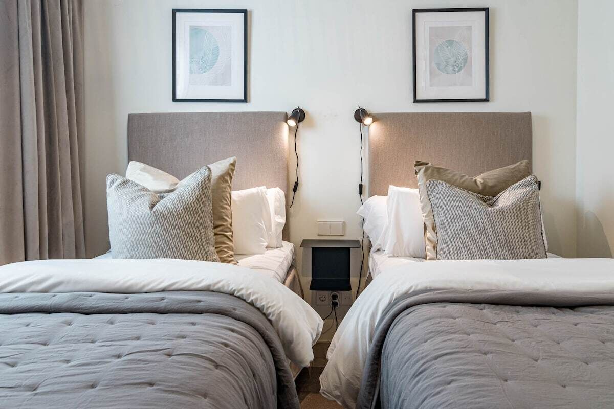 A hotel room with two beds, each with a headboard, bedside lamps, and a flat-screen TV.