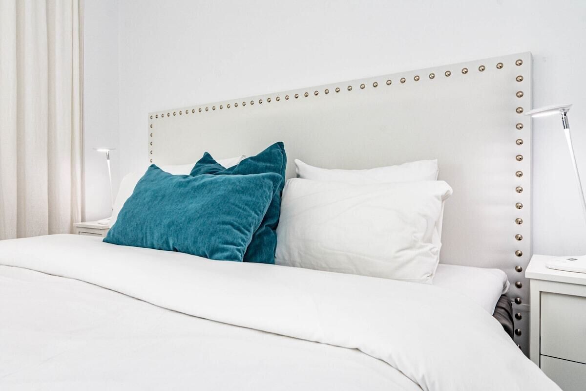 A neatly made bed with white and teal pillows, a white headboard with decorative studs, and a bedside table with a lamp.