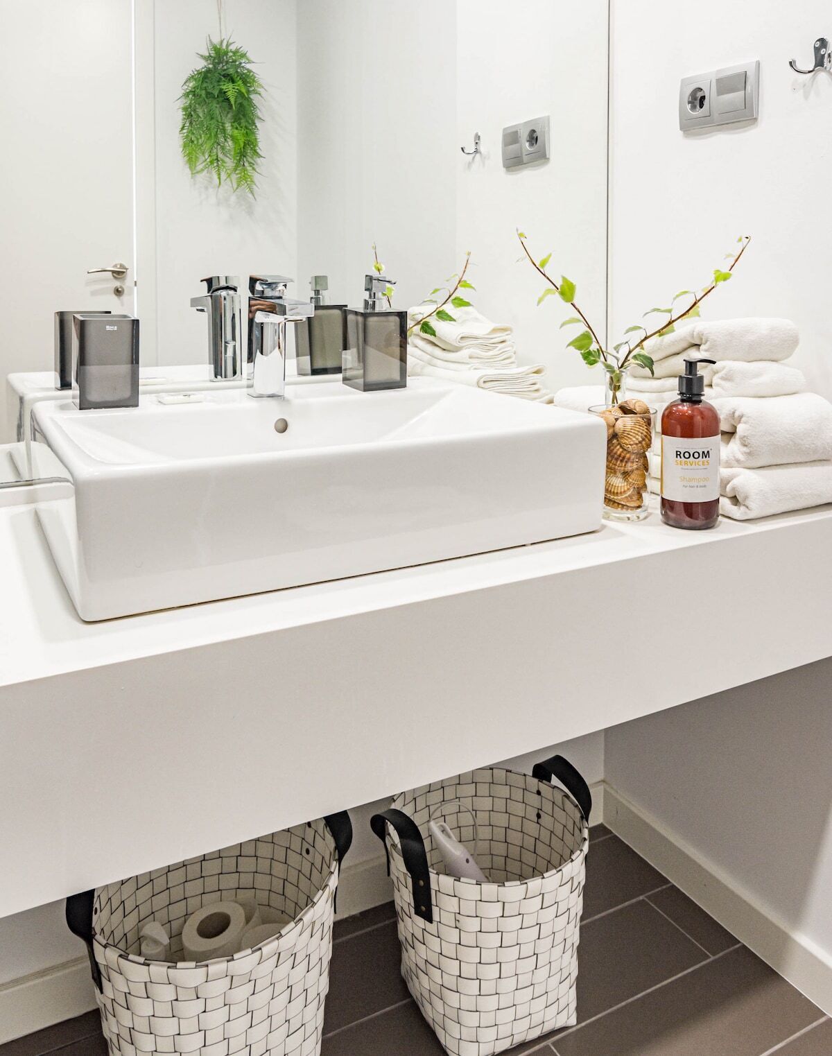Ein modernes Badezimmer mit einem großen weißen Waschbecken, mehreren Wasserhähnen, einem Spiegel, einer Pflanze, einer Shampooflasche und zwei gewebten Körben unter dem Waschbecken.