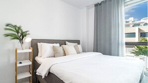 A modern bedroom with a large bed, a bedside table with a lamp, and a view of buildings through a window. - Lm111a- Modern Flat, Amazing Views (Estepona)