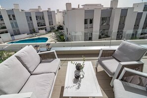 Una terraza en la azotea con piscina, mobiliario blanco y vistas a edificios modernos.