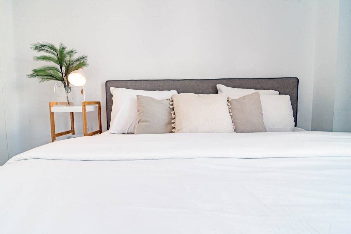 A neatly made bed with a grey headboard, white bedding, and a small bedside table with a lamp and a plant.