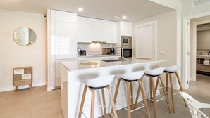 A modern kitchen with white cabinetry, a central island with stools, a microwave, and a built-in oven. - Lme2ba- Cozy Ground Floor Flat in Luxury Resort (Estepona)