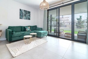 A modern living room with a green sofa, two round tables, and a large sliding glass door leading to an outdoor area. - Lme2ba- Cozy Ground Floor Flat in Luxury Resort (Estepona)
