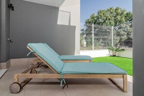 Two wooden lounge chairs with wheels, positioned on a tiled patio with a view of a fenced garden. - Lme2ba- Cozy Ground Floor Flat in Luxury Resort (Estepona)