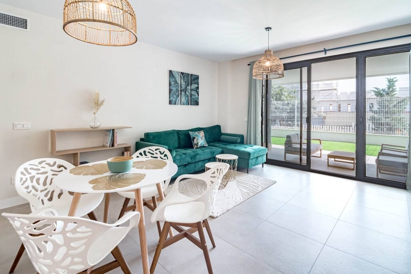 A modern living room with a green sofa, white chairs, a dining table, and a large sliding glass door leading to a balcony.