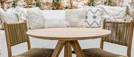 A round wooden table with four chairs, a stone wall background, and a bench with cushions.