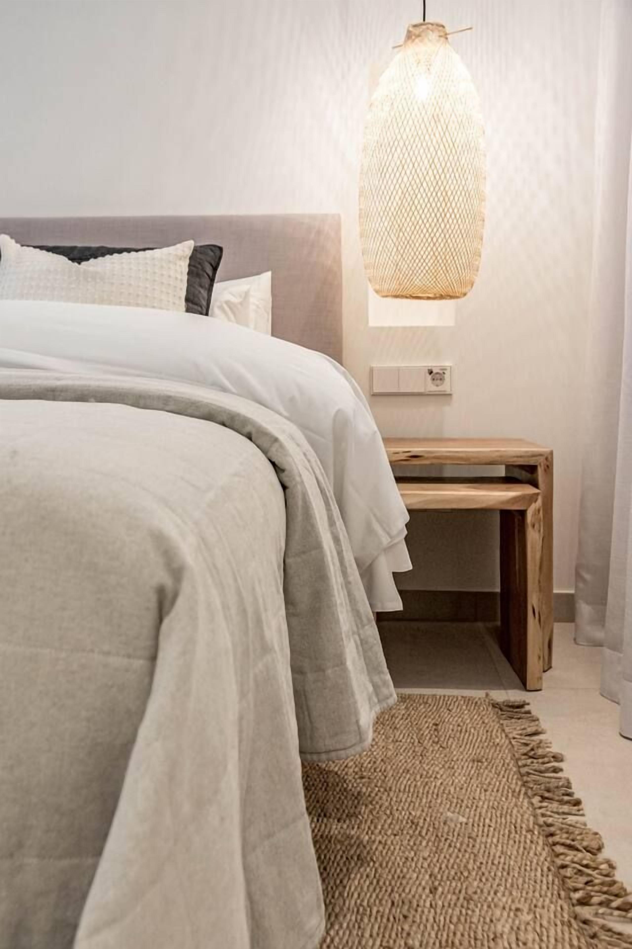 Una cama bien tendida con un edredón gris y almohadas, un tapete tejido, una mesita de noche de madera y una lámpara colgante.