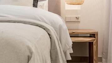 Cama bem arrumada com edredom cinza e travesseiros, tapete tecido, mesa de cabeceira de madeira e luminária pendente.