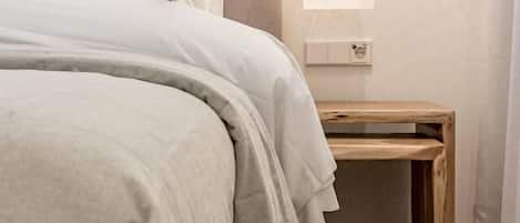 A neatly made bed with a grey comforter and pillows, a woven rug, a wooden nightstand, and a hanging light fixture.