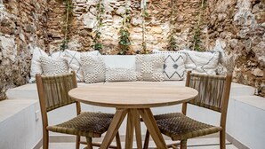 Una mesa redonda de madera con cuatro sillas tejidas, frente a una pared de piedra adornada con plantas colgantes.