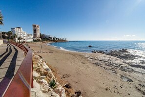 Ein Strand mit gepflastertem Weg, felsigem Ufer und Gebäuden im Hintergrund.