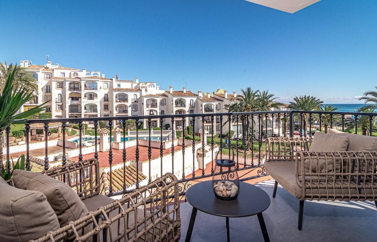 Een balkon met rieten meubilair, een ronde tafel en uitzicht op een zwembad en gebouwen.
