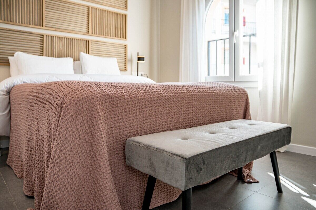 A neatly made bed with a pink textured blanket, a grey bench, and a wooden headboard.
