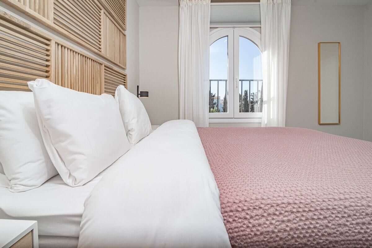 A bedroom with a large bed, white bedding, a pink blanket, a wooden headboard, a window with curtains, and a mirror on the wall.
