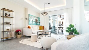 Un salon moderne avec un canapé blanc, une table basse et une bibliothèque. Une grande fenêtre offre une vue sur la ville.