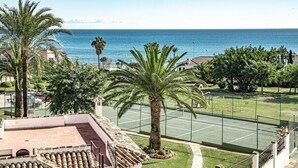 Un court de tennis offrant une vue sur la mer, entouré de palmiers et bordé d’un bâtiment doté d’un toit en tuiles.