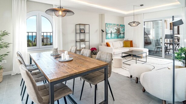 Un salon moderne avec un coin repas, comprenant une table en bois, des chaises à revêtement gris, un canapé blanc et une grande fenêtre donnant sur la mer.