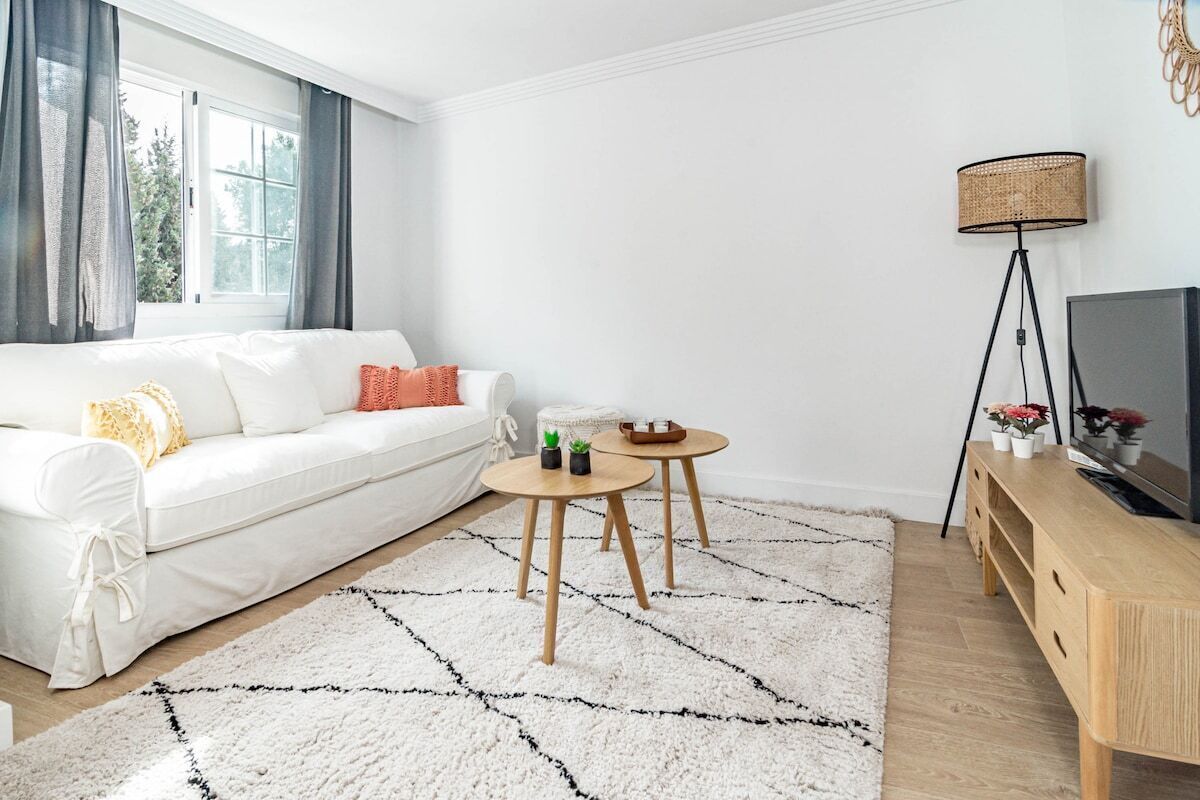 Ein modernes Wohnzimmer mit einem weißen Sofa, zwei kleinen Holztischen, einem weißen Teppich, einer Stehlampe und einem Fernsehschrank.