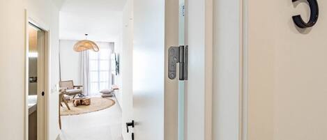 A hallway with a door marked with the number 5, a light switch, and a view into a room with a bed, a chair, and a hanging light fixture.