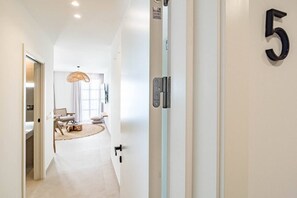 Un pasillo con una puerta marcada con el número 5, un interruptor de luz y vistas a una habitación que tiene una cama, una silla y una lámpara colgante.