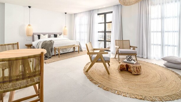 Una habitación de hotel moderna con una cama grande, dos mesitas de noche, un tapete redondo tejido, una mesa de comedor con sillas y un ventanal con cortinas.
